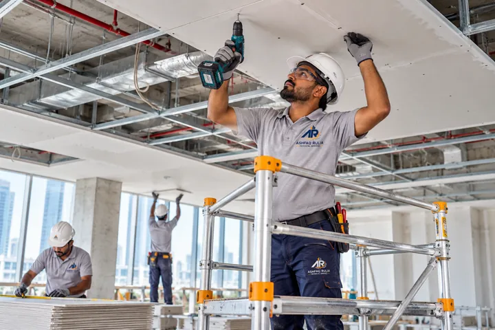 An ARTS ceiling installer fitting a gypsum false-ceiling panel from a proper low mobile scaffold with guardrails, UAE commercial interior